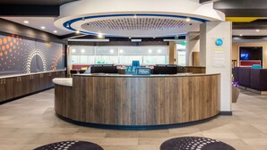 Ein moderner Hotel-Empfangsbereich mit einer gebogenen Holztresen, einem digitalen Bildschirm und Blick ins Hotel-Lobby.