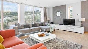 A modern living room with a grey sofa, a red armchair, a coffee table with snacks, a TV, and a large window offering a city view. - Close To The Beach,Wi-Fi, Parking (Challaborough)