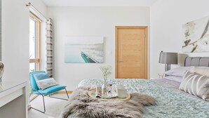 A modern bedroom with a large bed, a wooden door, a window with curtains, a painting on the wall, and a bedside table with a lamp. - Close To The Beach,Wi-Fi, Parking (Challaborough)