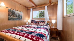 A cozy bedroom with a wooden bed, a painting of a cabin, and a window with curtains. - Finch Nest - Fireplace, Deck, Walk to Lake & Village (Big Bear Lake)