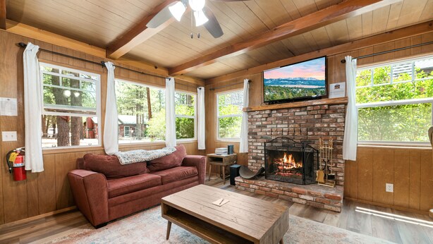 A cozy living room with a brick fireplace, wooden ceiling, and large windows. - Finch Nest - Fireplace, Deck, Walk to Lake & Village (Big Bear Lake)