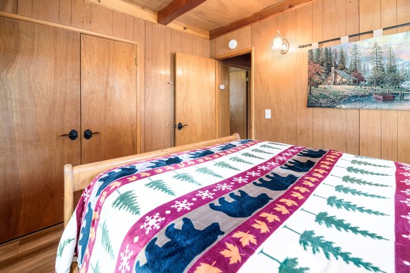 Een kamer met houten lambrisering, een bed, een schilderij van een hut en een deur naar een andere ruimte.