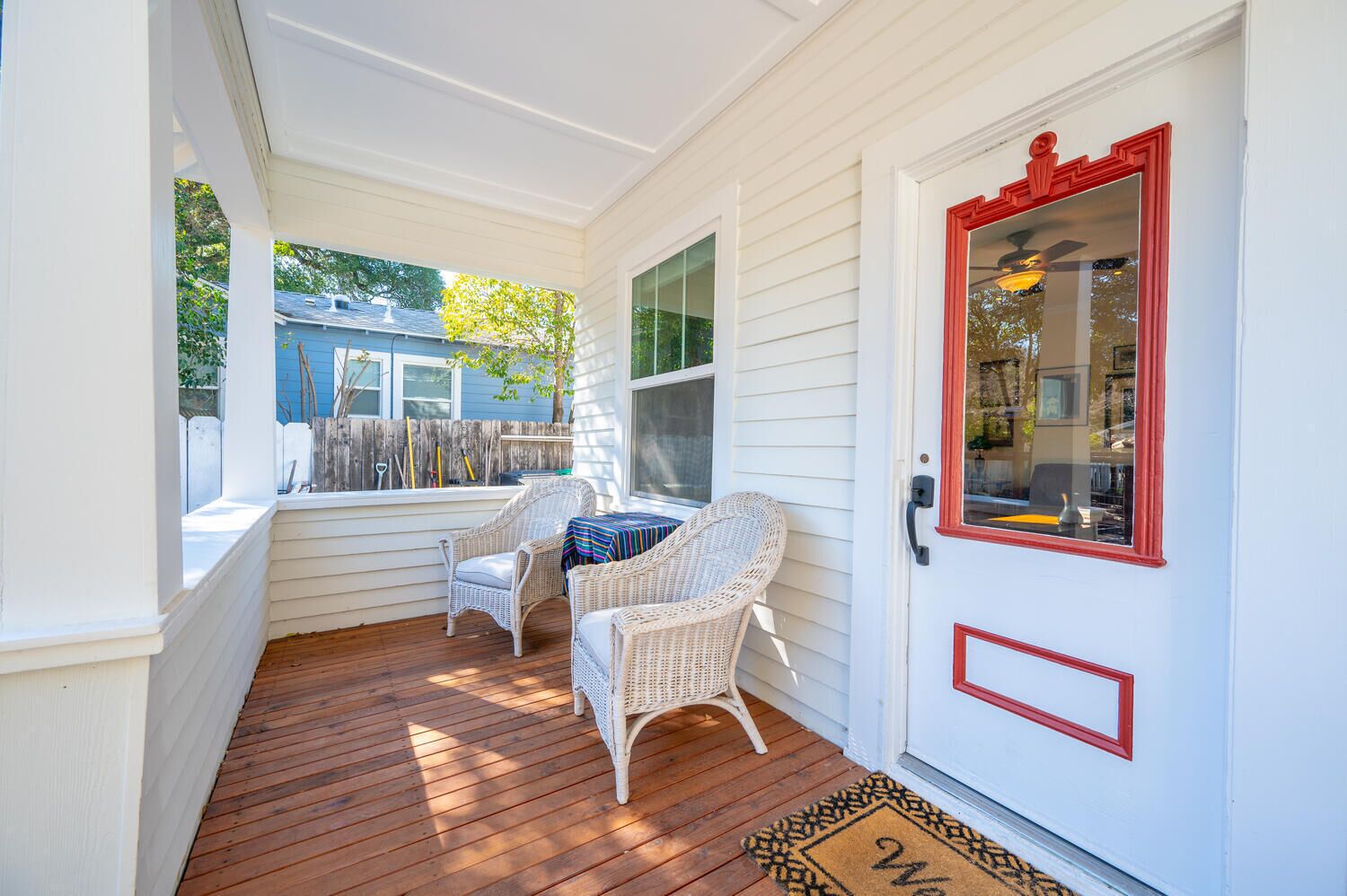 A porch with wicker chairs and a small table, a wooden deck, and a white door with a red frame.