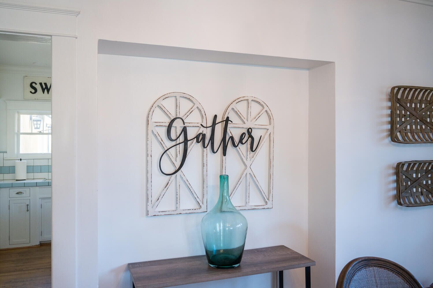 A wall with a 'Gather' sign and a decorative window frame. A teal vase is on a wooden table.