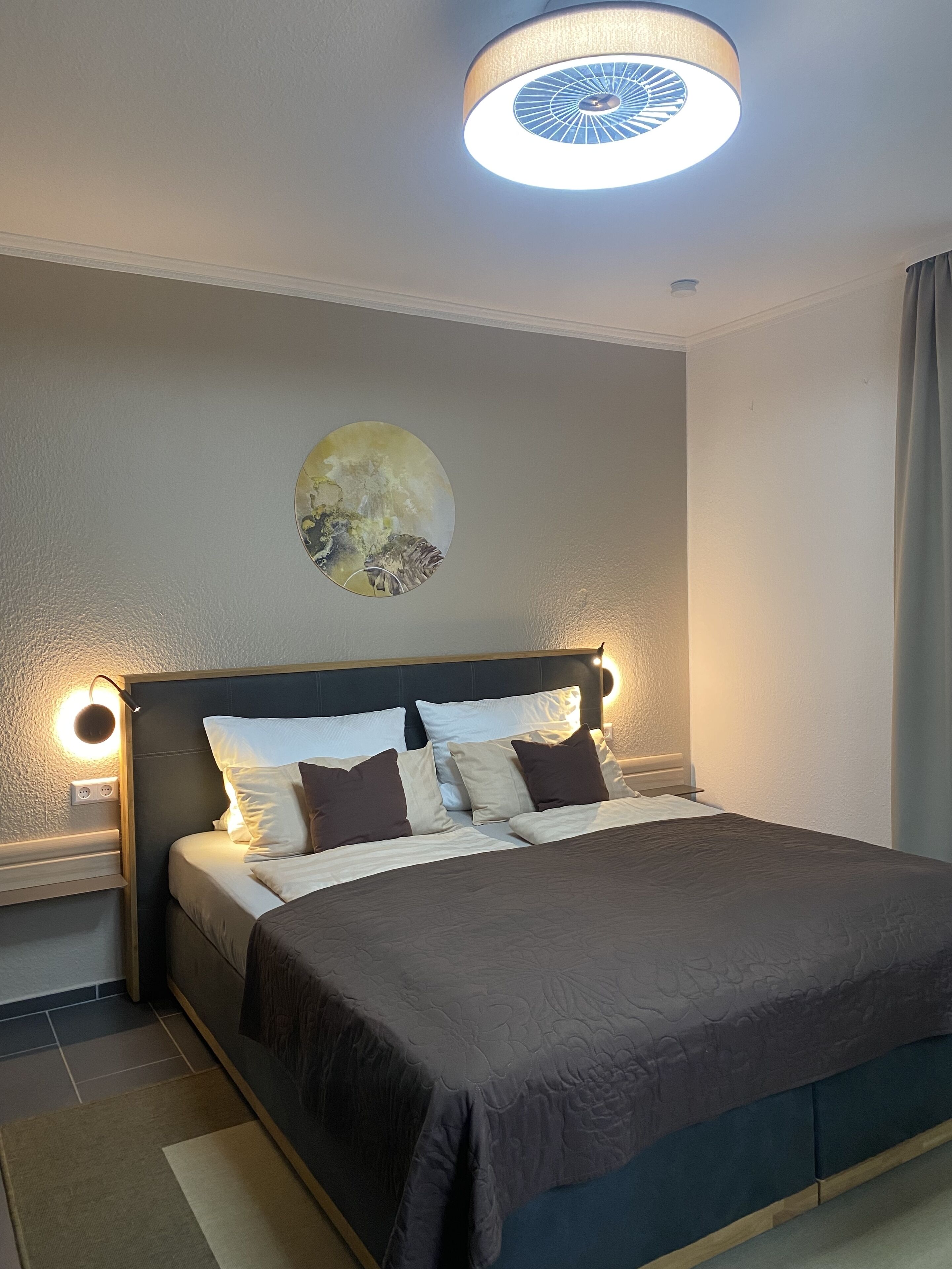 A neatly made bed with a dark headboard, a wall-mounted light fixture, and a circular ceiling light.