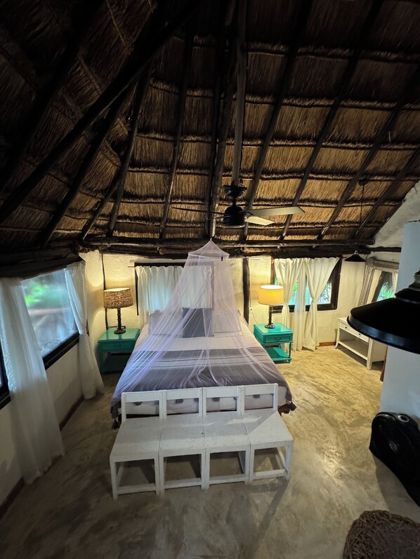 Una habitación con una cama bajo un techo de paja, mosquitera y ventilador de techo.