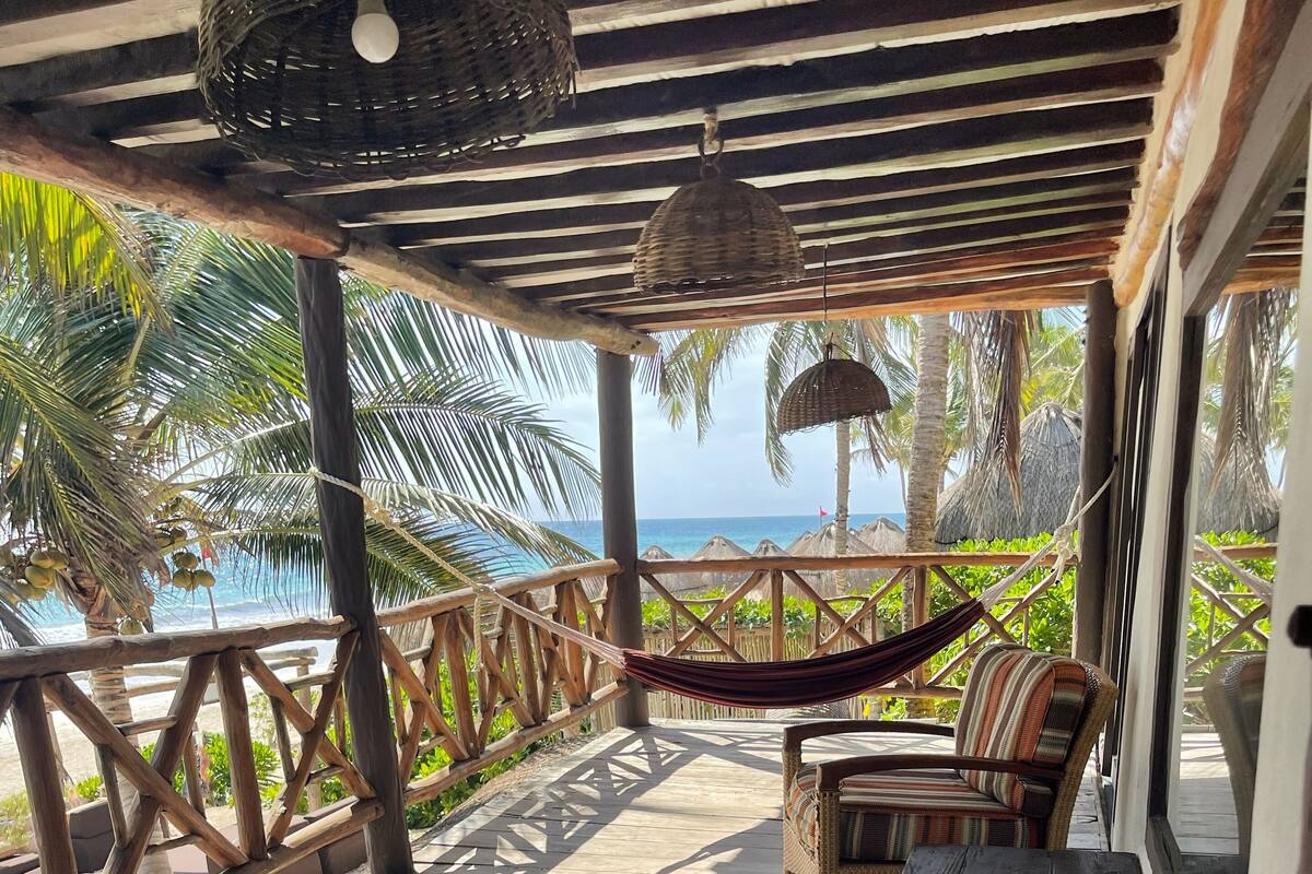 A covered outdoor seating area with wicker furniture and a hammock, overlooking a beach and palm trees.