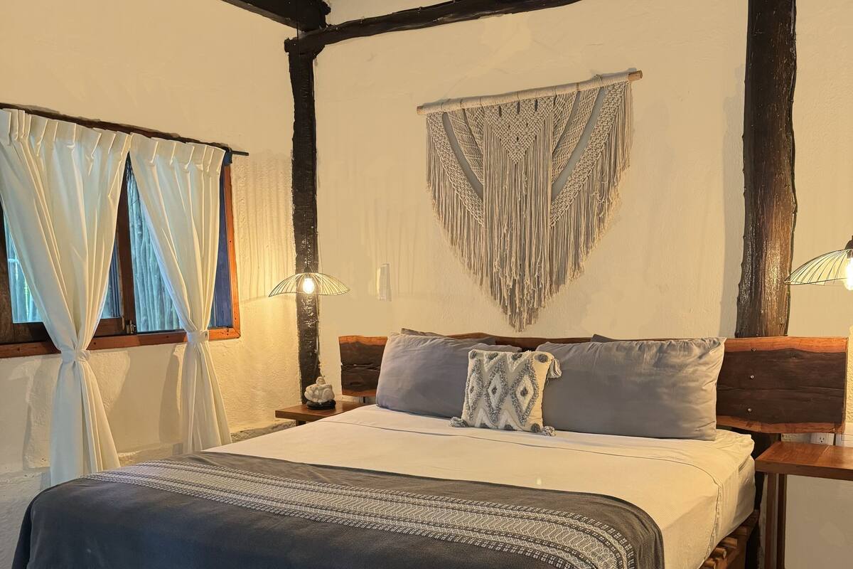 A bedroom with a bed, bedside tables, a bench, a macrame wall hanging, and curtains.