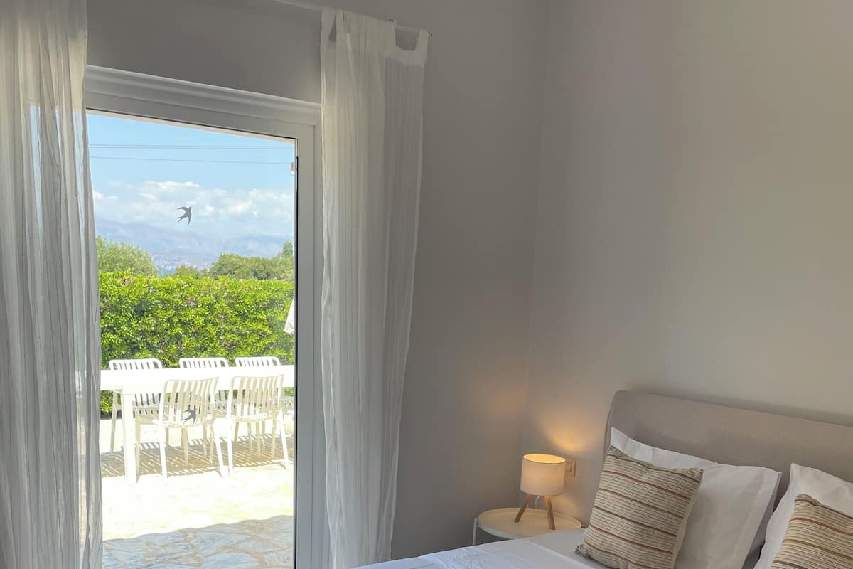 A bedroom with a bed, white curtains, a nightstand with a lamp, and a view of an outdoor area with chairs and greenery.