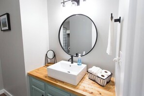 Un baño con un lavamanos blanco, un espejo redondo, una encimera de madera, una cesta pequeña con toallas y un cuadro en la pared.
