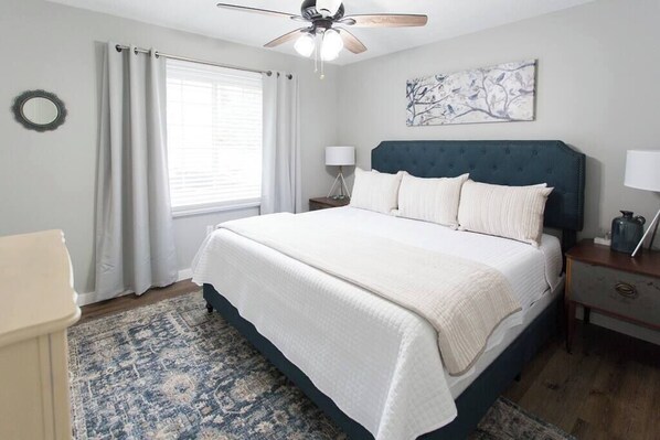 A bedroom with a bed, a nightstand, a lamp, a ceiling fan, a window with curtains, and a wall art piece.