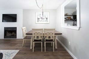 Un comedor con una mesa y sillas de madera, una chimenea, un televisor montado en la pared y una cocina visible a través de una ventana.