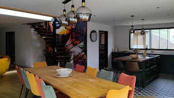 Un comedor moderno con una mesa de madera y sillas coloridas, una escalera con un mural de colores y una cocina con un ventanal grande.