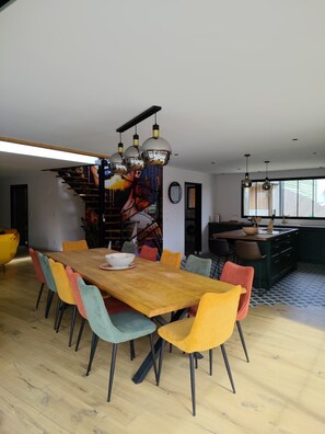 Un espace repas moderne avec une table en bois et des chaises colorées, un escalier orné d’une fresque colorée et une cuisine dotée d’une grande fenêtre.