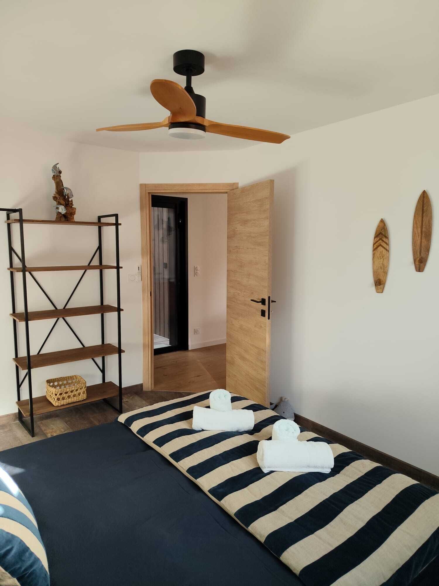 A bedroom with a bed, a wooden shelf, a ceiling fan, and a door leading to another room.