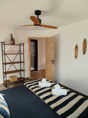 Un dormitorio con cama, repisa de madera, ventilador de techo y puerta que da a otra habitación.