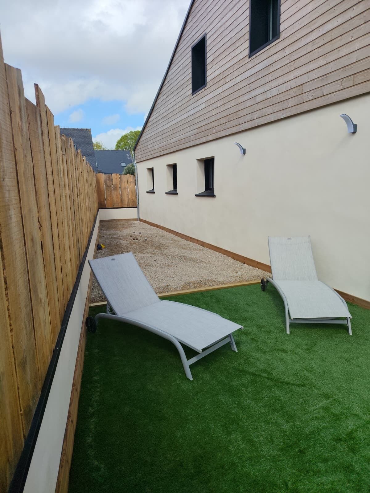 Un patio con césped artificial, dos sillones de relax, una valla de madera y un edificio con ventanas.