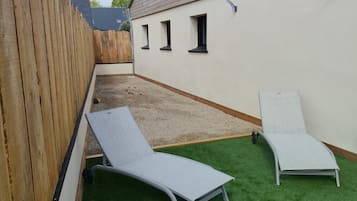 Un patio con césped artificial, dos sillones de relax, una valla de madera y un edificio con ventanas.