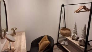 Una habitación con una silla negra, una mesita de madera, un espejo, una cesta colgante y una estantería con zapatos y objetos decorativos.