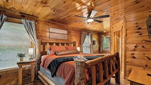 A wooden cabin bedroom with a bed, a nightstand, a ceiling fan, and a window with curtains.