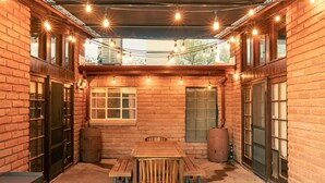 Un patio couvert avec une table et des bancs en bois, des murs en briques et des guirlandes lumineuses.