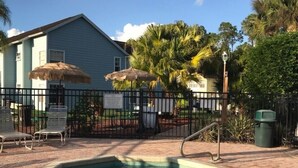 A swimming pool with a brick border, surrounded by lounge chairs and umbrellas, with a house and palm trees in the background. - Recently Remodeled, Very Comfy Condo Near Disney, Universal and Sea World. (Kissimmee)