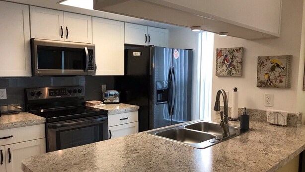 A modern kitchen with white cabinets, a stainless steel microwave, a black refrigerator, a granite countertop, and a double sink. - Recently Remodeled, Very Comfy Condo Near Disney, Universal and Sea World. (Kissimmee)