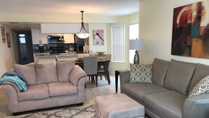 A living room with a beige sofa, a grey sofa, and a patterned ottoman. There is a dining area with a table and chairs in the background. - Recently Remodeled, Very Comfy Condo Near Disney, Universal and Sea World. (Kissimmee)