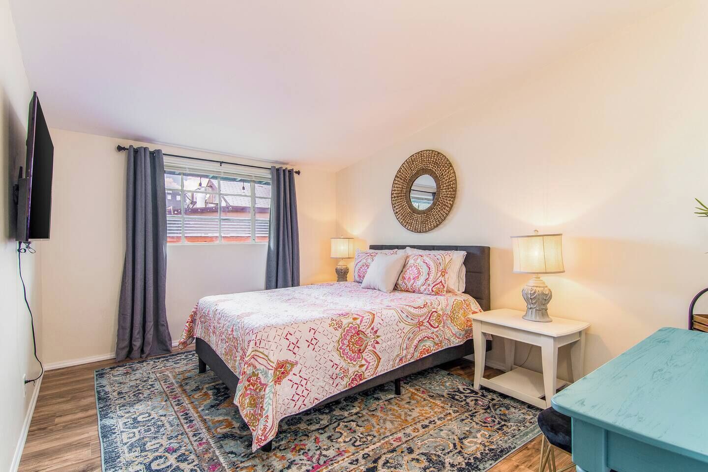 Une chambre à coucher avec un lit, deux lampes de chevet, une télévision, une fenêtre avec des rideaux et un tapis à motifs.