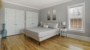 A bedroom with a bed, two white doors, a window with blinds, and a nightstand with a lamp.