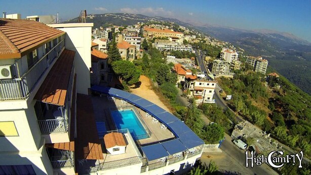 A high-rise building with a rooftop swimming pool, surrounded by a hilly townscape. - Hotel Le Crillon (Broumana)