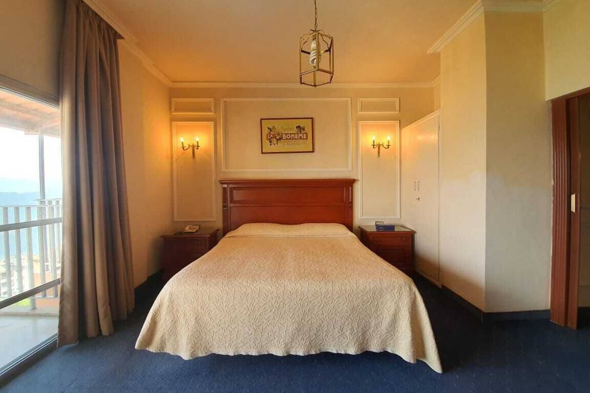 A hotel room with a large bed, two bedside tables, a nightstand, a chandelier, a framed picture on the wall, and a view of the outdoors through the window.