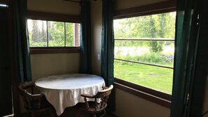 Ein Zimmer mit einem runden Tisch, der mit einer weißen Tischdecke bedeckt ist, zwei Holzstühlen und Blick auf eine grüne Grünfläche durch große Fenster mit grünen Vorhängen.