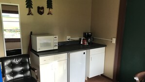 Eine kompakte Küchenzeile mit Mikrowelle, Kaffeemaschine und kleinem Kühlschrank. Es gibt einen schwarzen Ledersessel und einen Holzsessel. Der Raum hat eine Tür mit Blick nach außen.