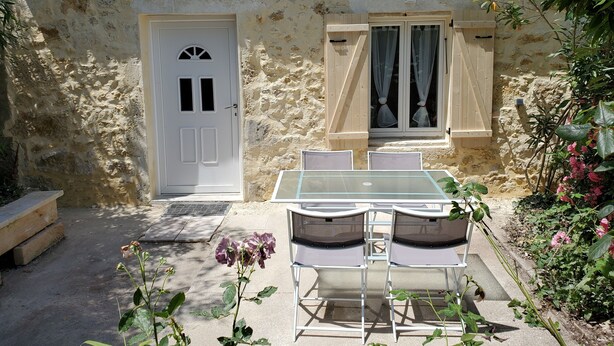 A rustic stone building with a tiled roof, a white door, and a window with curtains. There is a table with chairs in front, and a bench to the left.