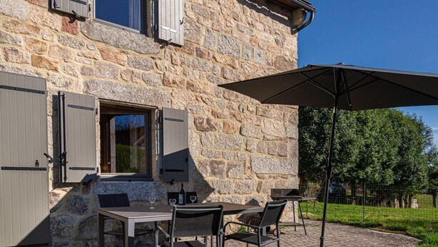 A stone building with a patio area featuring a table and chairs, a large umbrella, and a wine bottle.