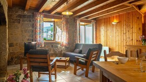 A cozy room with a stone wall, wooden ceiling beams, a sofa, wooden chairs, a table, and a window with curtains.