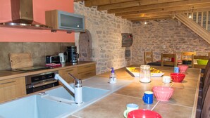 A kitchen with a stone wall, wooden cabinets, a sink, and a dining area with colorful bowls. - Gîte "La Prairie" - LA CANOURGUE (LA CANOURGUE)