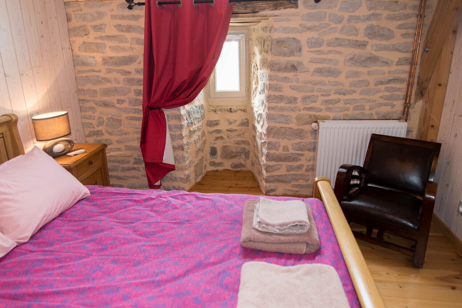 A bedroom with a stone wall, a bed with a pink and blue patterned bedspread, a wooden nightstand with a lamp, a chair, and a window with a red curtain.