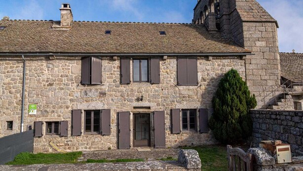A stone building with a steep roof, multiple windows with shutters, and a small door.