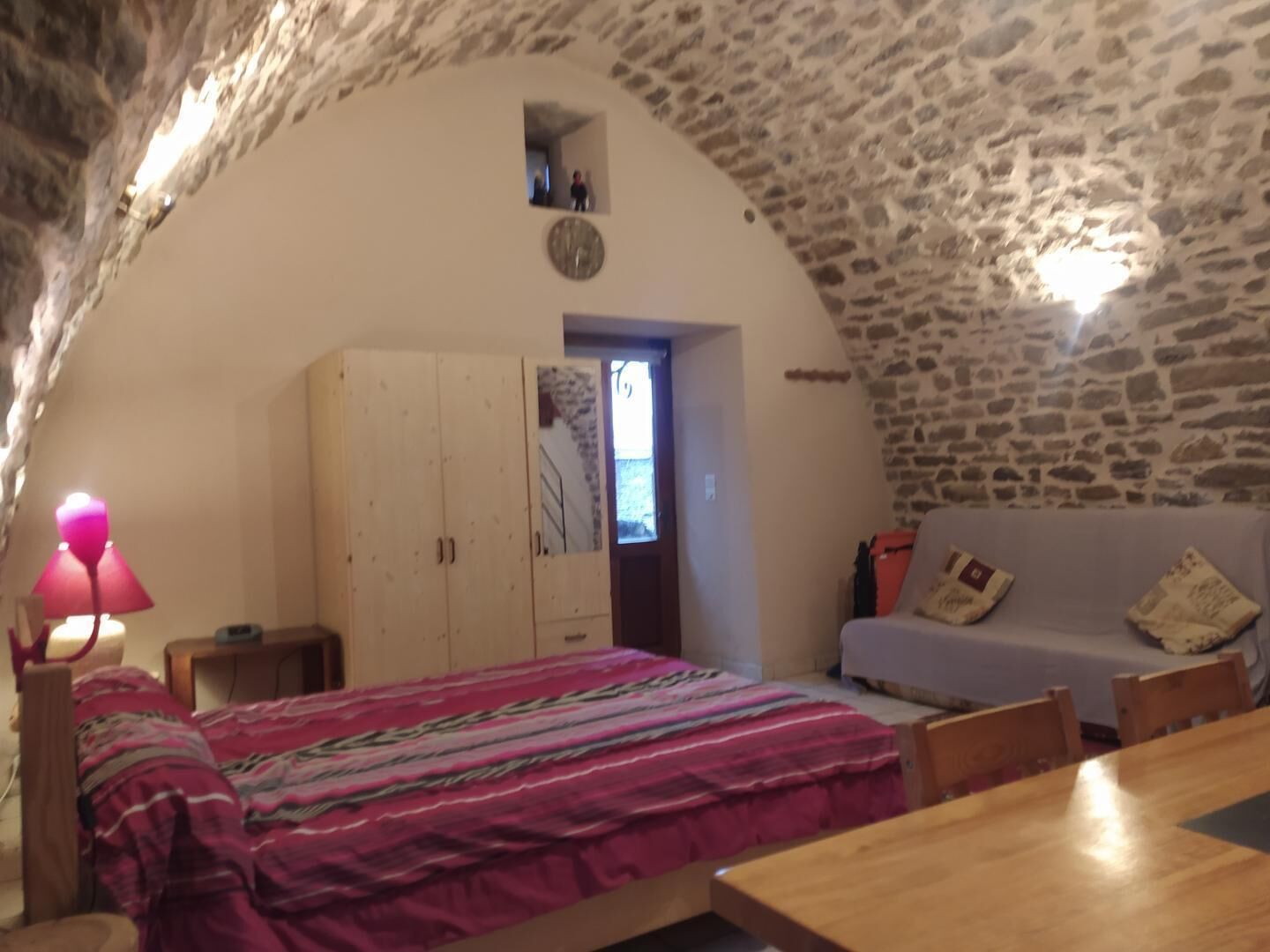 Une pièce chaleureuse avec un plafond en pierre, un lit recouvert d’une couverture colorée, une table en bois, un canapé et une petite fenêtre.