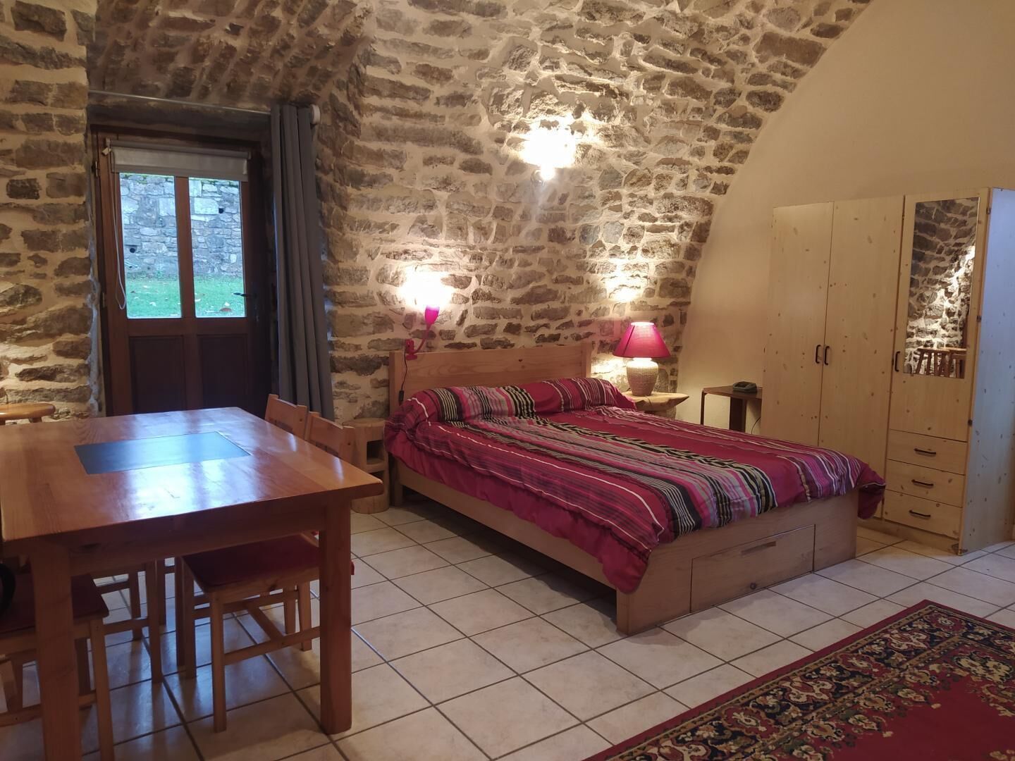 A bedroom with a stone wall, a wooden bed, a nightstand with a lamp, a wardrobe, and a dining table with chairs.