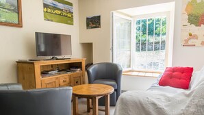 A cozy living room with a TV, wooden cabinet, leather chair, round table, and a bed with a red pillow.