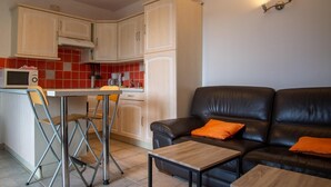Un espace de vie compact comprenant un coin cuisine, un coin repas avec une petite table et des chaises, et un coin salon avec un canapé et deux petites tables d’appoint.