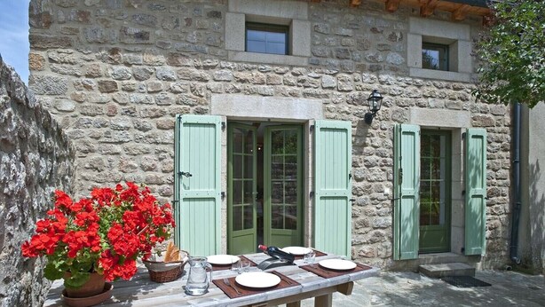 Un edificio de piedra con contraventanas verdes, una mesa dispuesta para comer y una maceta con flores rojas.