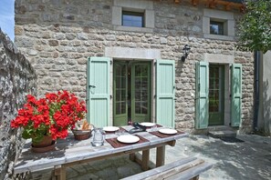 A stone building with green shutters, a table set for dining, and a potted plant with red flowers. - Gîte Le Mas 6 persons (ALLENC)