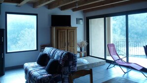 A room with a wooden ceiling, a sofa, a table, a cabinet, and a balcony with a chair.
