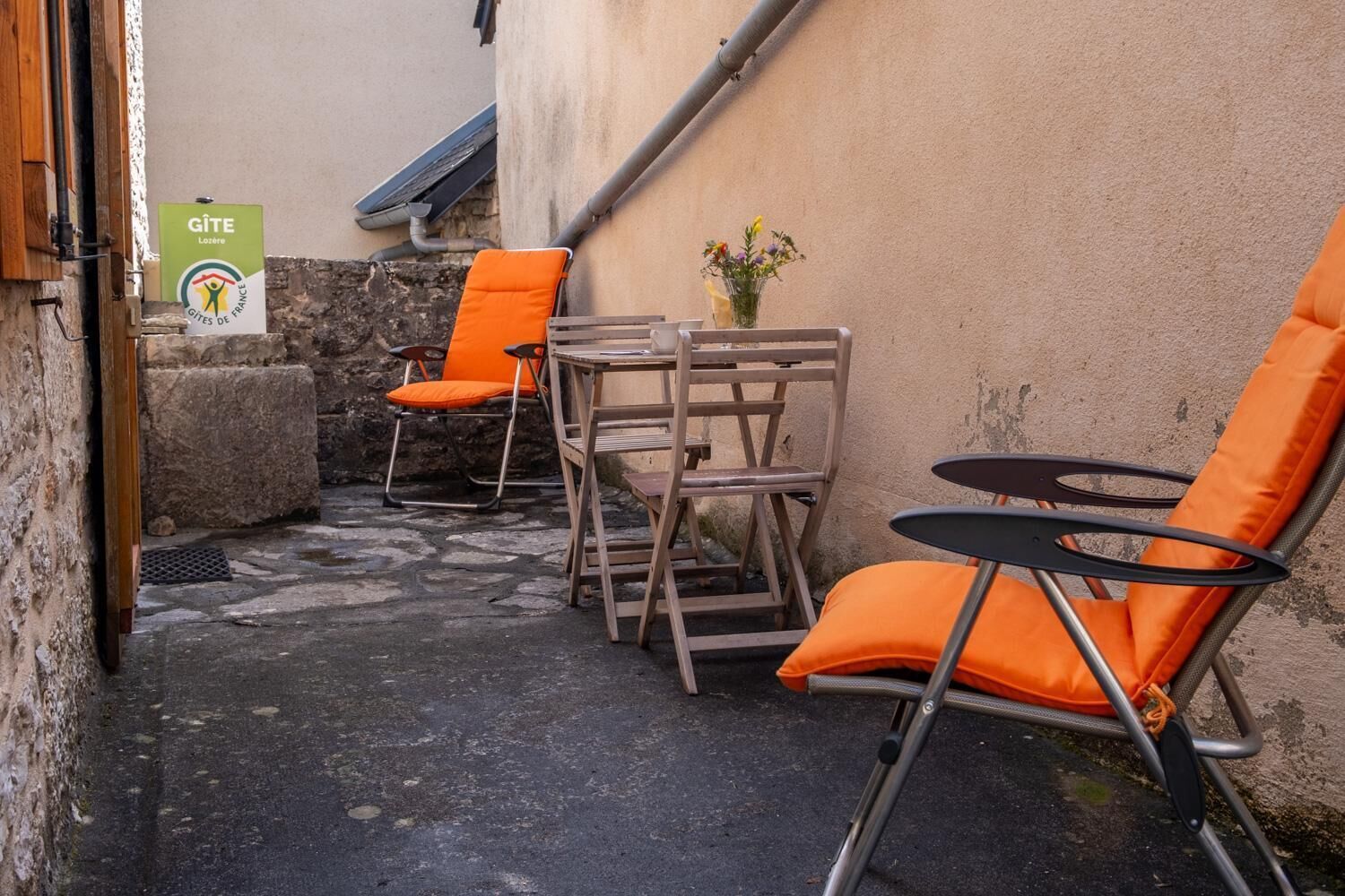 Een terras met oranje stoelen en een tafel, een stenen muur en een bord met Gîte Lozère erop.