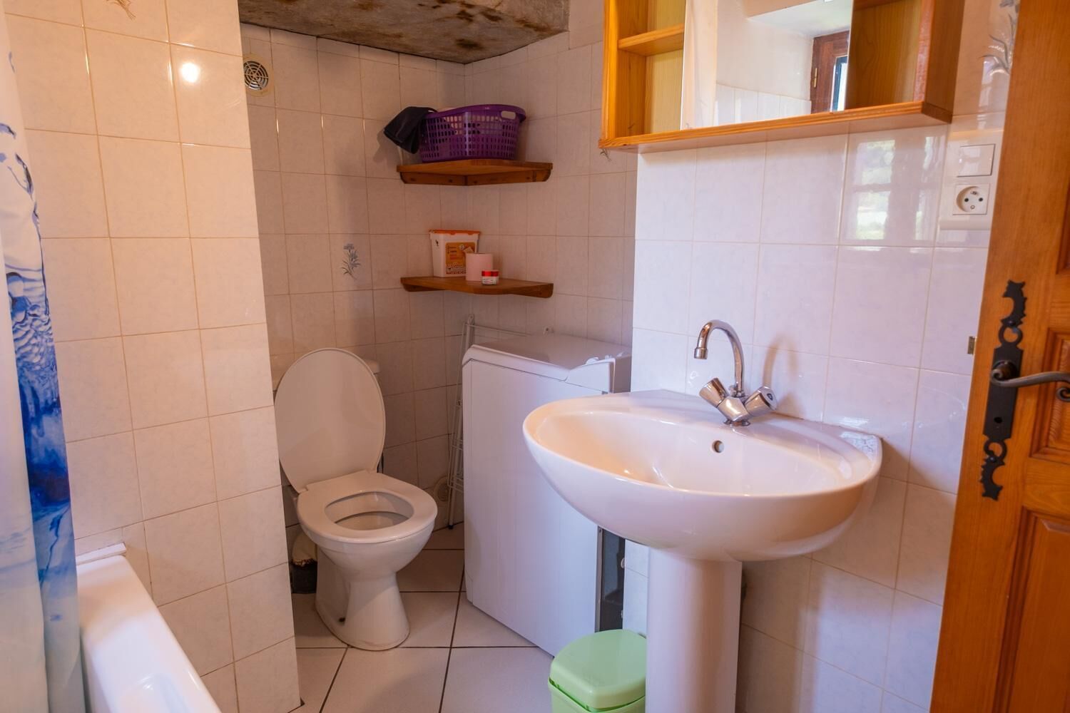 Een kleine badkamer met toilet, wastafel en douche. Boven het toilet bevindt zich een plank en boven de wastafel een spiegel.