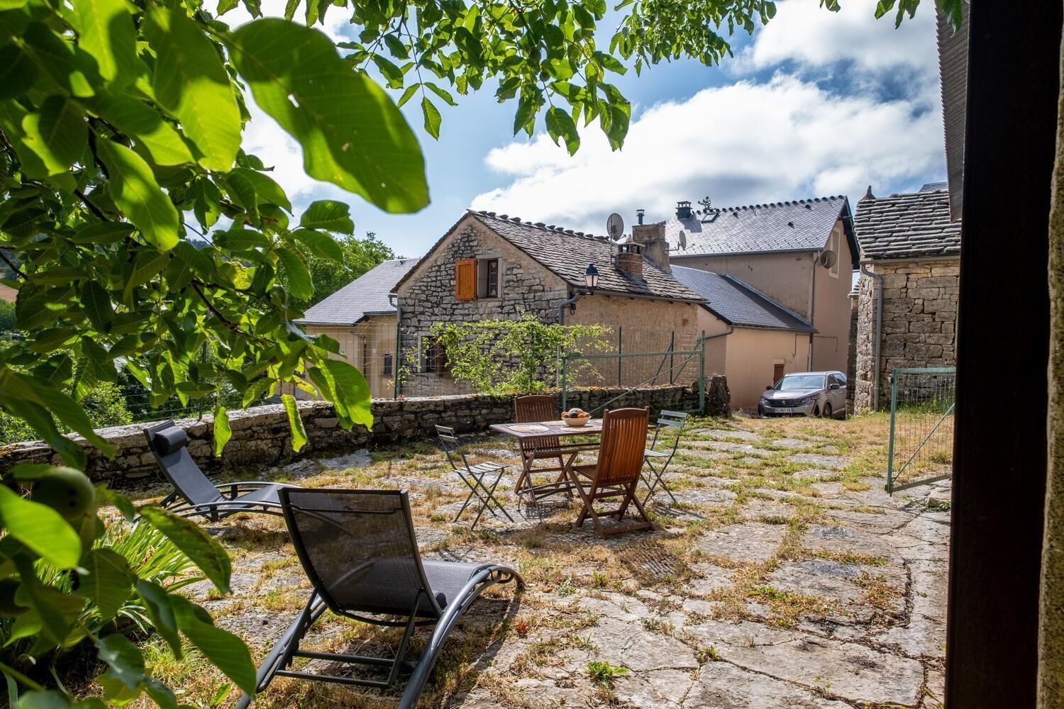 Une terrasse en pierre avec des meubles d’extérieur, un bâtiment en pierre avec un toit en tuiles et une voiture garée à proximité.